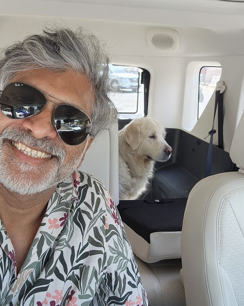 Thommen Jose smiling in aviator sunglasses and a floral shirt, with a golden retriever sitting behind him in the car
