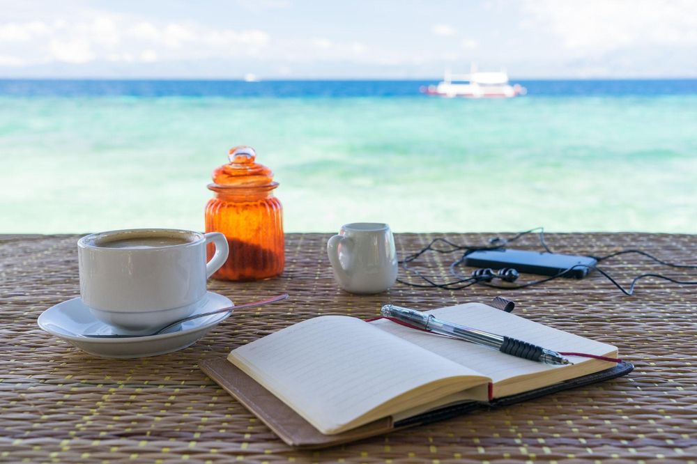 A coffee cup and an open notebook with a pen on a table beside a turquoise sea