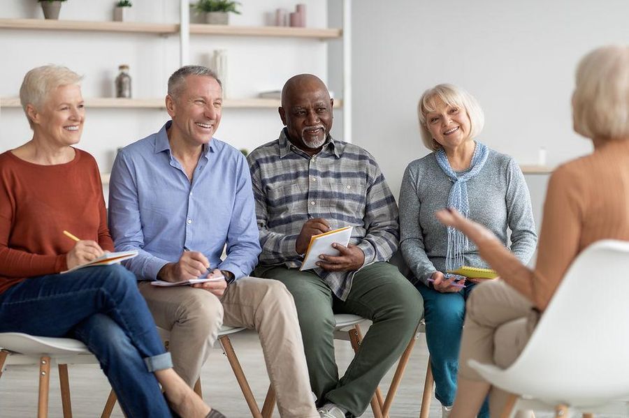 A relaxed circle of mature adults smiling and taking notes as they listen to a speaker