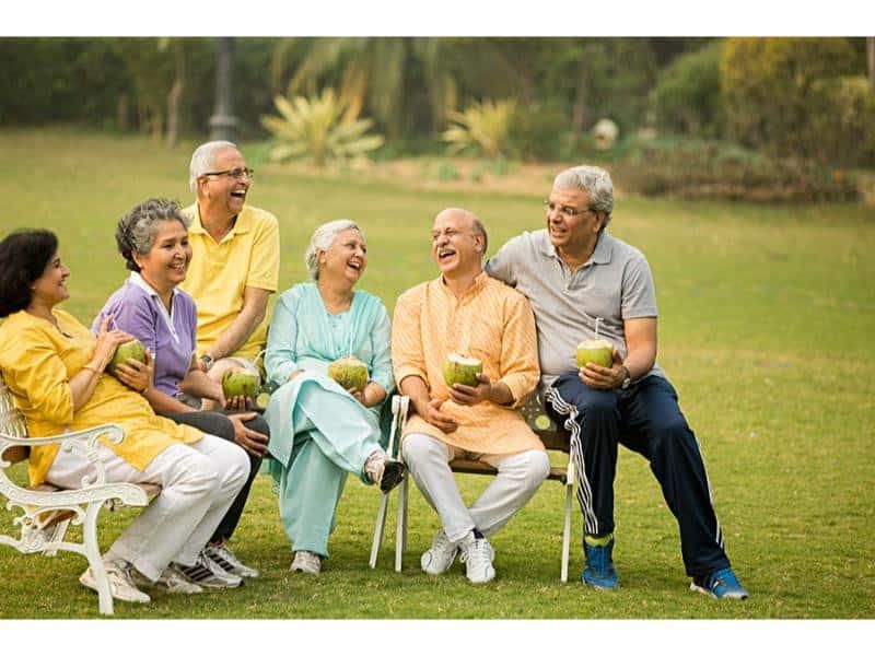 Six friends in their fifties and sixties laughing together on a bench in a park, holding tender coconuts