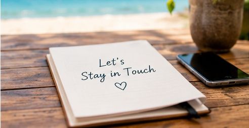 An open notebook on a wooden table reading Let's Stay in Touch, with a phone and a plant beside a sunlit beach
